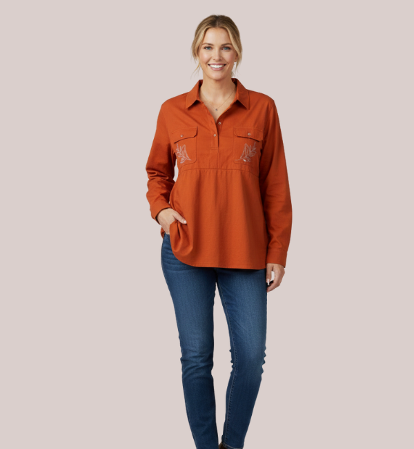 Woman in a burnt orange long-sleeve customizable breastfeeding shirt, featuring a quarter-button placket, twin chest pockets, and white custom leaf-patterned embroidery on the chest.