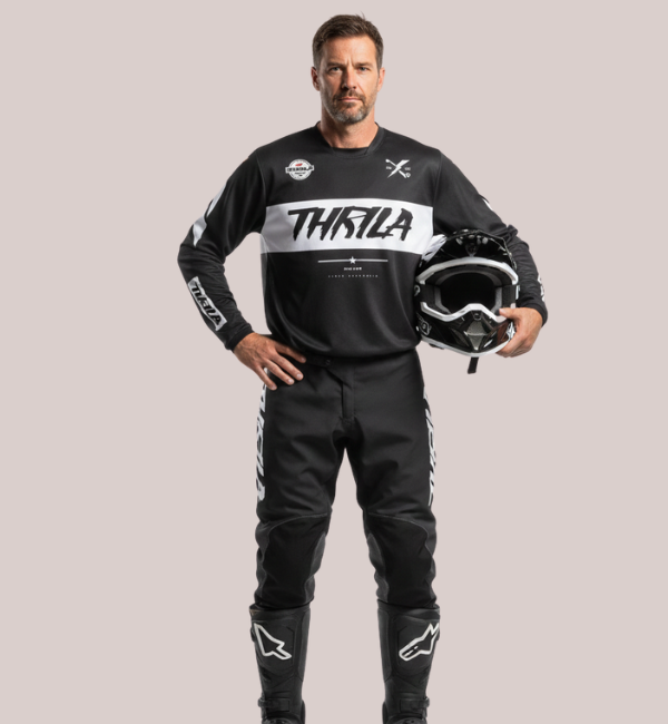Man wearing a black and white Thrila Pro-Series long-sleeve motocross jersey and pants, customizable racewear kit, holding a black dirt bike helmet.